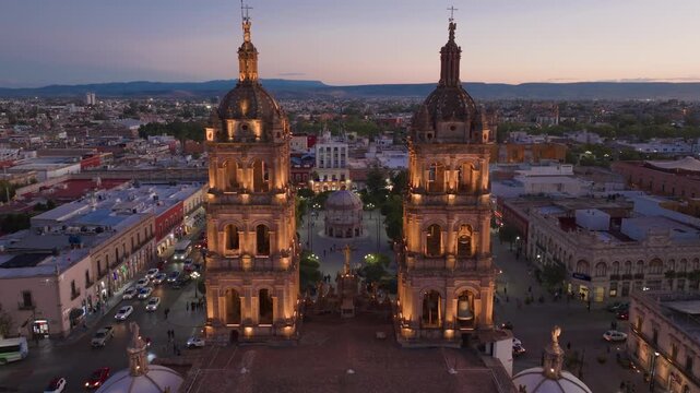 Aerial drone footage of Cathedral Basilica in Durango Mexico at twilight with golden illuminated twin towers, pink sunset sky, mountains in distance, and bustling city plaza below.
