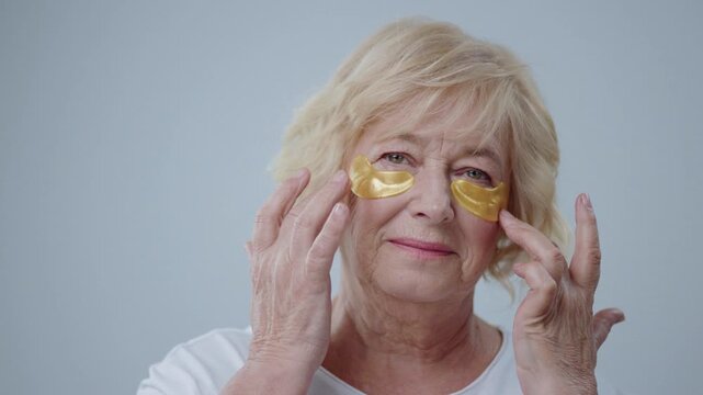 Joyful mature woman with lovely smile wearing eye patches on face. Portrait of senior lady doing everyday facial cleaning routine in the bathroom.