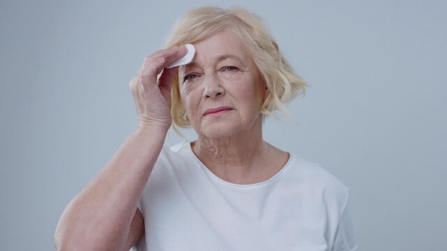 Appealing blonde woman in white shirt clearing her face with lotion looking in the mirror against grey background. Portrait of nice-looking grandma doing her everyday beauty routine at home.
