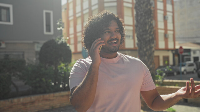 Man holding smartphone to ear and gesturing with hand on a sunny street near a building; casual chat contentment.