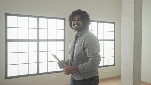 Indian man holding a clipboard and writing with pen, right hand visible, standing in an empty building with large windows; confidence property sale.