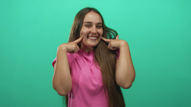 Woman pressing cheeks with index fingers on green studio set while puffing cheeks and smiling slightly; playful cheeky pose.