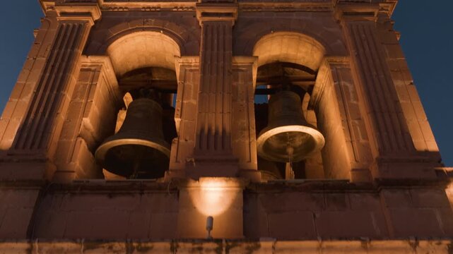Historic cathedral bell tower and dome illuminated by warm golden lights against deep blue twilight sky in Durango Mexico, featuring ornate baroque architecture with twin bells and clock face.