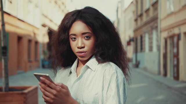 Attractive young Afro-American woman actively uses her smartphone, then seductively looks straight to camera. Model, beauty, posing to camera, romantic atmosphere. Female portrait