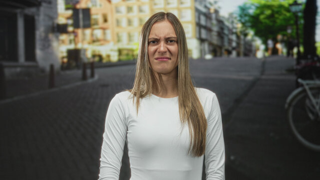 Young hispanic woman grimaces with scrunched nose and pursed lips on a street, standing center frame; annoyance.