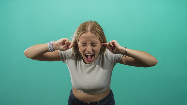 Woman covers ears with fingers and screams in turquoise studio wearing white crop top and jeans; frustration release.
