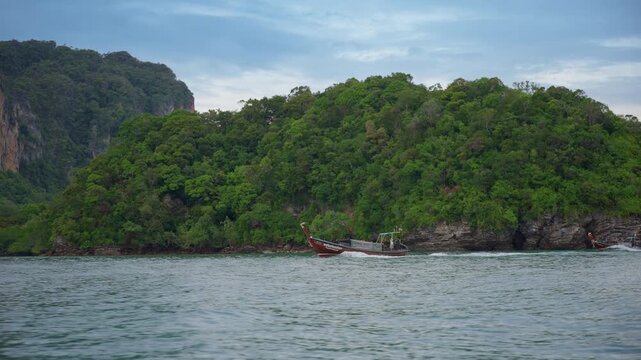 Traditional Thai longtail boats cruise through turquoise waters in slow motion near Mueang Krabi Thailand. Lush green jungle covers dramatic limestone cliffs rising from Andaman Sea under cloudy.