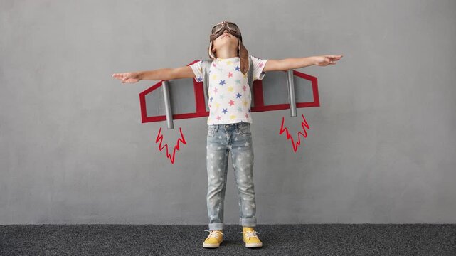 Happy child playing with paper rocket against grey wall background. Kid having fun indoors. Funny boy pretend to be astronaut. Imagination and children dream concept