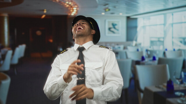 Young hispanic pilot smiles and tilts head, wearing captain hat, uniform shirt, epaulettes and tie in airport lounge; confidence welcome.