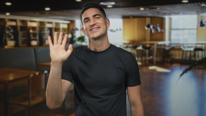 Obraz premium Man waving hand in building lobby with bare forearm visible and wearing black t shirt while smiling to camera; friendly greeting.