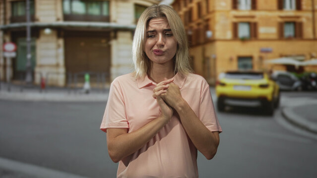 Woman clasping hands in a pleading gesture on a street with buildings and a yellow car, pink polo shirt visible; uncertainty plea.
