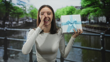 Fototapeta premium Young woman holding a wrapped gift box with blue ribbon and cupping her hand to mouth on a street by a canal railing, smiling broadly; surprise joy celebration.