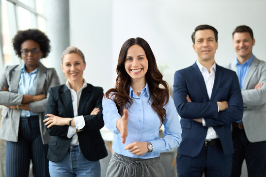 Happy young businesswoman offering a handshake in the office while looking at camera. Her colleagues are in the background.