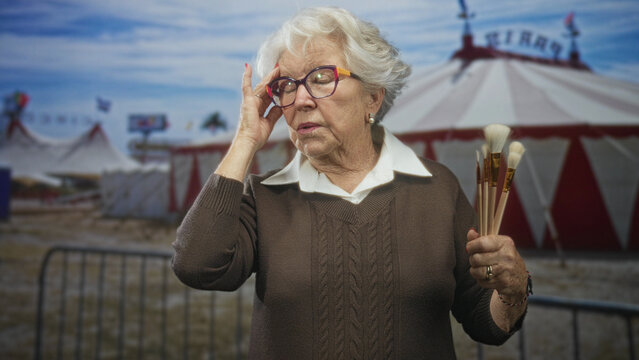 Elderly woman holding paintbrushes touches eyeglasses on street in front of circus tent, slight frown and thoughtful gaze; nostalgia calm.