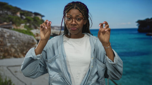 Woman wearing glasses and denim shirt holding hands in air quotes gesture at studio seaside set with turquoise ocean backdrop; skepticism irony.