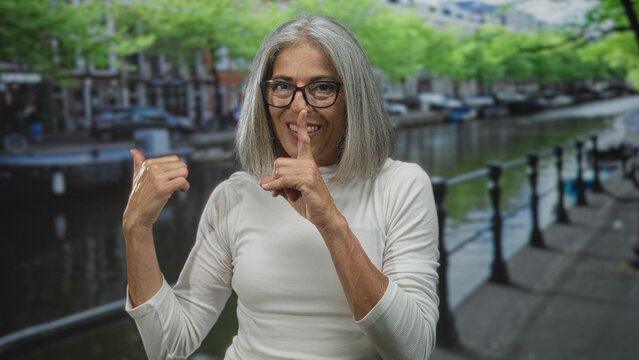 Woman with finger to lips for silence on an amsterdam canal street, wearing glasses and smiling while thumb points to herself; playful secret.