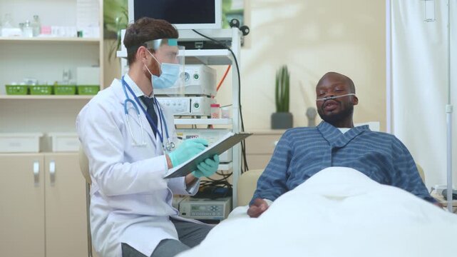 Adult doctor in full-face profection shield mask talking with sick patient during coronavirus quarantine at hospital. Doctor appointment. Doctor checkup.