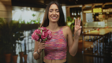 Fototapeta premium Woman holding pink bouquet and holding up two fingers while smiling in restaurant interior; celebration joy.
