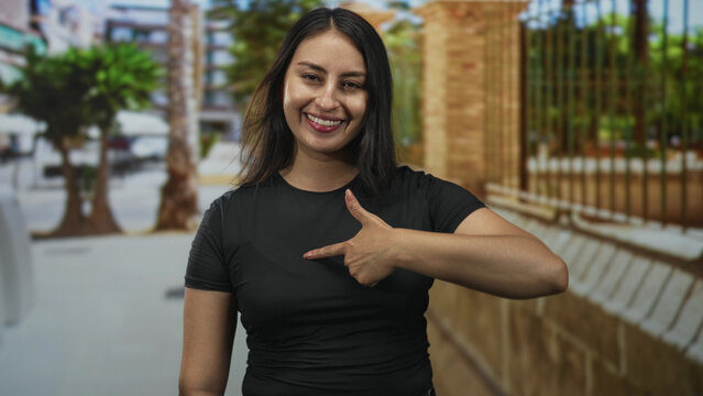 Young hispanic woman smiling and giving thumbs up while pointing with her right hand on a sunny street with blurred trees and brick fence; warm happiness.