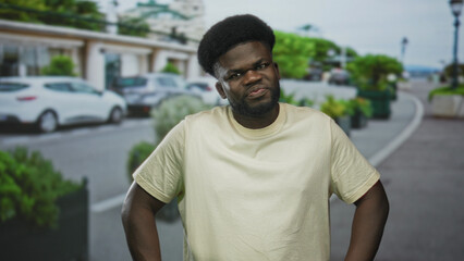 Fototapeta premium Man with hands on hips and frowning expression wearing beige tshirt on a city street; frustration defiance.
