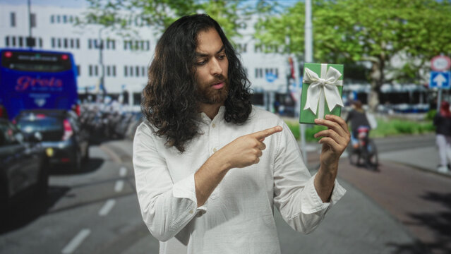 Man pointing at a green gift box on a busy city street, holding it up amid traffic and cyclists while studying the ribbon; curiosity gift discovery.