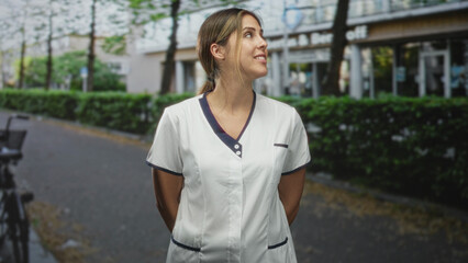 Obraz premium Woman nurse in white scrub uniform standing with hands behind back on street, smiling; friendly compassion.