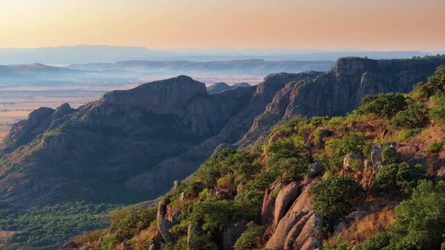 Breathtaking aerial view of rugged mountain cliffs covered in green vegetation during golden hour sunset, with dramatic rocky formations extending into misty valleys and warm orange sky in background.