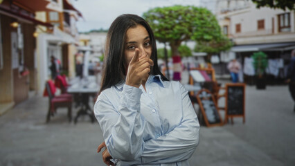 Fototapeta premium Woman points finger to cheek with arms crossed on a restaurant terrace near sidewalk tables, facing camera and smiling slightly; playful teasing confidence.