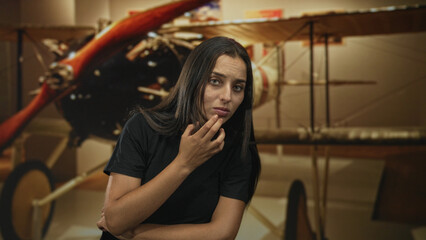 Young hispanic woman, finger to lips beside a vintage biplane in a museum building with exhibit lighting and aircraft propeller visible  contemplation. © Krakenimages.com