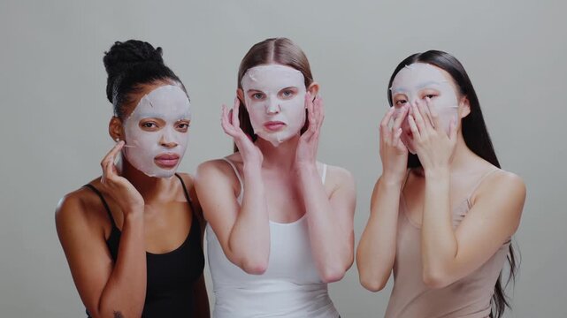 Beauty treatment. Multi-ethnic skincare models wearing purifying sheet masks massaging their faces enjoying cosmetic product advertising on background.
