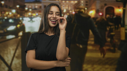 Fototapeta premium Woman smiling with arms crossed on a lit city street at night beside iron railing and blurred traffic lights; confidence ease.