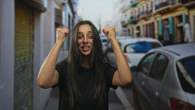 Hispanic woman smiling with fists raised and clenched hands, bare forearms visible, standing among parked cars on a narrow city street with blurred buildings; joy victory.