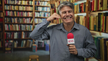 Man holding a red news microphone and smiling while making a phone gesture to his ear among bookshelves in a library building  playful banter. © Krakenimages.com