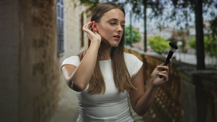 Fototapeta premium Young woman holds a makeup brush and touches her ear while checking her face beside a stone balustrade on a tree lined street; confidence beauty routine.