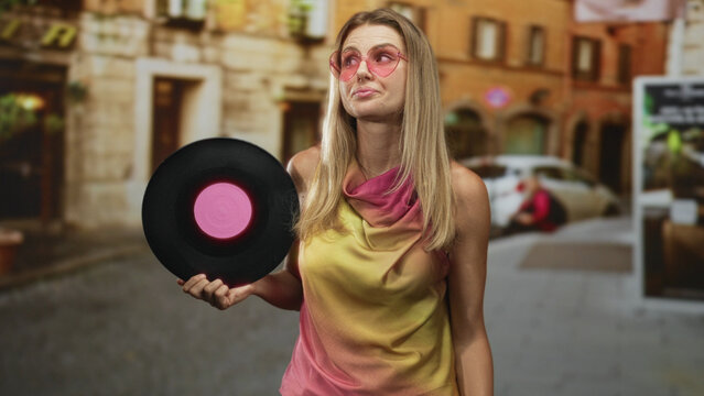 Woman holding vinyl record with hand on head and pink sunglasses on a cobbled street; nostalgia retro.