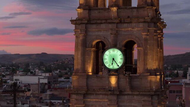 Cathedral Basilica of Immaculate Conception in Durango, Mexico features illuminated bell tower with glowing green clock face. Baroque stone architecture against pink and purple sunset sky over city.