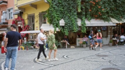 Obraz premium Busy cobblestone street scene softly blurred with shallow bokeh, defocused cafe terrace and hanging ivy on building facade street; background backdrop copyspace.