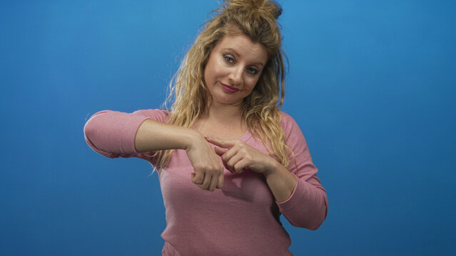 Woman blonde points finger to wrist and taps hand while looking at her palm in a blue studio; impatience waiting.