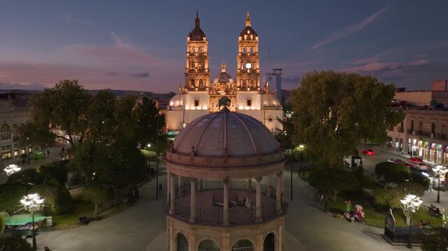 Cathedral Basilica of Immaculate Conception in Durango, Mexico illuminated at dusk. Aerial view captures ornate baroque twin towers, central dome, and historic plaza with gazebo and trees at blue.