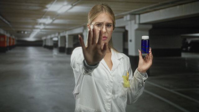 Woman in white coat holding blue pill bottle, palm raised in stop gesture inside parking garage; refusal caution.