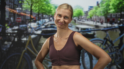 Fototapeta premium Woman smiling with bare midriff and hands on hips in a street scene with bicycles and canal; confidence wellness energy.