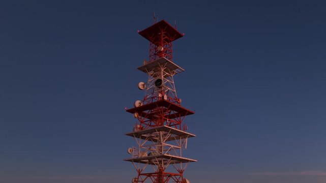 Tall telecommunications tower equipped with multiple satellite dishes and red warning lights stands against a gradient twilight sky transitioning from deep blue to soft orange and purple hues.