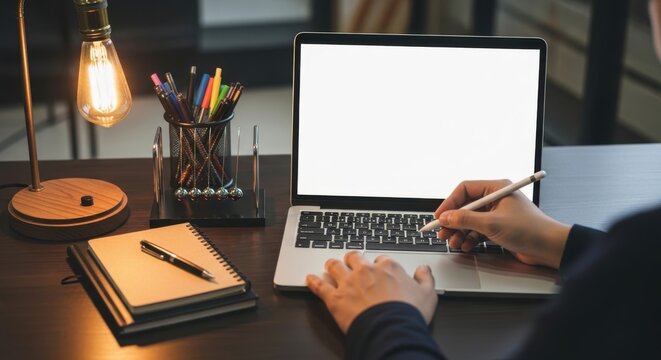 Person working on laptop at desk with notebook, pens, and vintage lamp, office setup