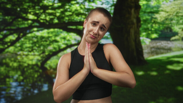 Woman with buzz cut pressing hands together at chest in forest by a pond, pleading expression and upward gaze; hopeful plea.