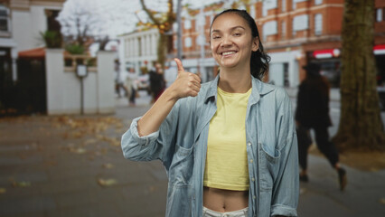 Fototapeta premium Young woman hitchhiking with thumb out on a busy street in front of brick buildings, wearing cropped yellow top and denim shirt; friendly optimism.