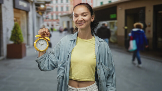 Woman holds yellow alarm clock by the handle at chest height, smiling and showing bare midriff on city street; urgency time awareness.