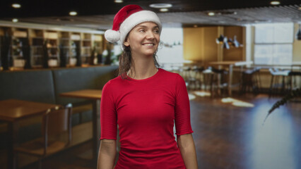 Obraz premium Woman wearing santa hat smiling and looking up inside a building cafe with wooden tables and chairs; cheerful holiday.