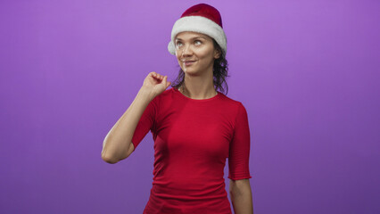 Obraz premium Young hispanic woman in santa hat with finger to chin and subtle smirk in purple studio; holiday cheer playful.