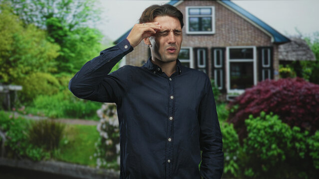 Man presses forehead with hand while squinting in front of a suburban building garden, wearing a blue shirt and earring; headache distress.