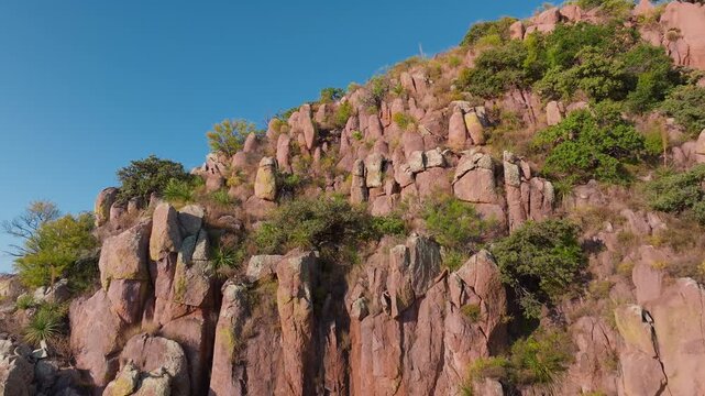 Aerial drone footage captures stunning pink granite rock formations dotted with yucca plants and desert shrubs across the rugged Sierra Madre Occidental mountains under clear blue skies in Mexico.